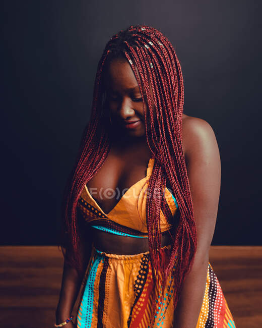 Cheerful African American female with pink braids standing on black background in studio and looking down — Stock Photo