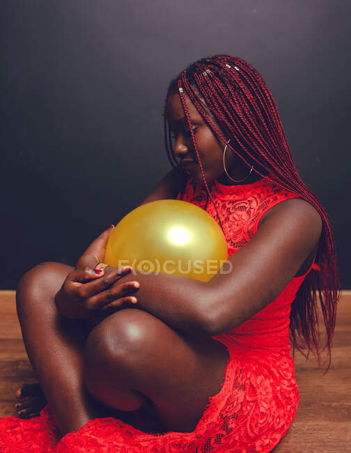 Vista lateral de la hembra afroamericana en vestido rojo sentada en el suelo con globo aerostático en estudio sobre fondo oscuro - foto de stock
