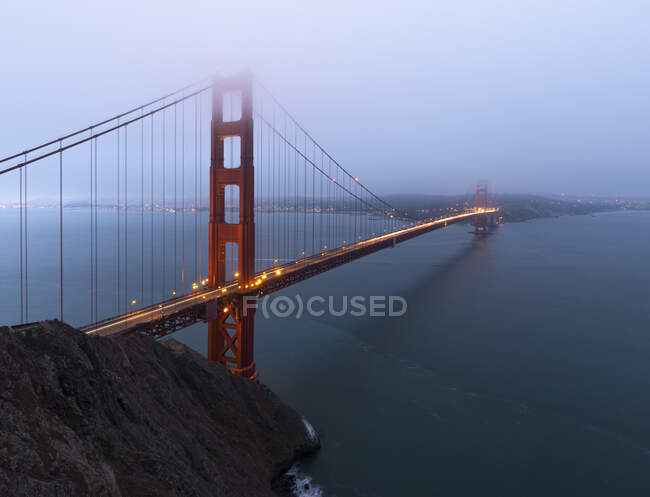 Золоті ворота міст з сяючим світлом над спокійним морем проти прибережного міста покриті туманом у вечірній час у Сан-Франциско. — стокове фото