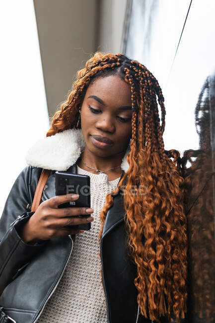 D'en bas Afro-Américaine en tenue tendance et avec des tresses appuyées sur un mur de verre du bâtiment et un smartphone de navigation en ville — Photo de stock