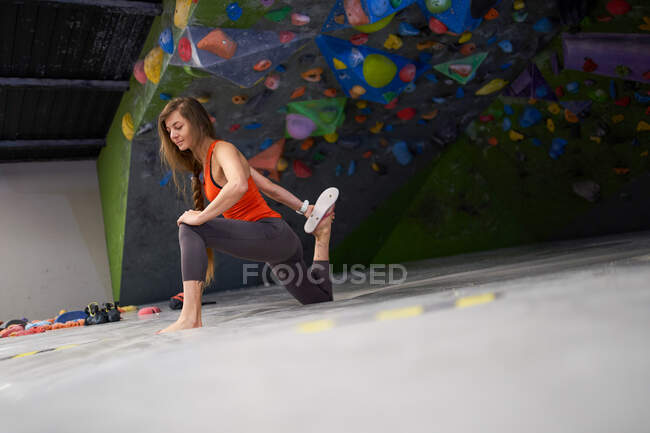 Volle Länge der jungen fokussierten Bergsteigerin in aktiver Kleidung, die Beine streckt, während sie im Boulderzentrum trainiert — Stockfoto
