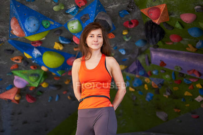 Encantadora mujer fuerte en ropa deportiva cogida de la mano detrás de la espalda mientras está de pie contra la pared de escalada y mirando a la cámara - foto de stock