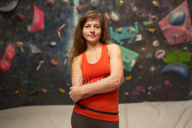 Encantadora mujer fuerte en ropa deportiva con brazos cruzados de pie cerca de la pared de escalada y mirando a la cámara - foto de stock
