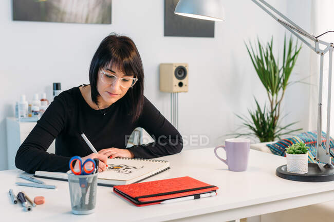 Artista feminina talentosa focada desenhando no bloco de notas enquanto se senta à mesa na sala de estar em casa — Fotografia de Stock