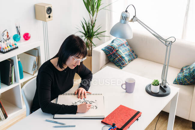 Von oben eine talentierte Künstlerin, die im Notizblock zeichnet, während sie zu Hause am Tisch im Wohnzimmer sitzt — Stockfoto