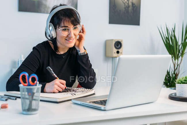 Lächelnde Künstlerin zeichnet Skizzenbuch, während sie mit Kopfhörern am Tisch sitzt und zu Hause Online-Unterricht auf Netbook schaut — Stockfoto