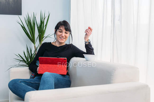Felice femmina seduta sul divano e guardando film divertenti su tablet mentre si rilassa nel fine settimana a casa — Foto stock