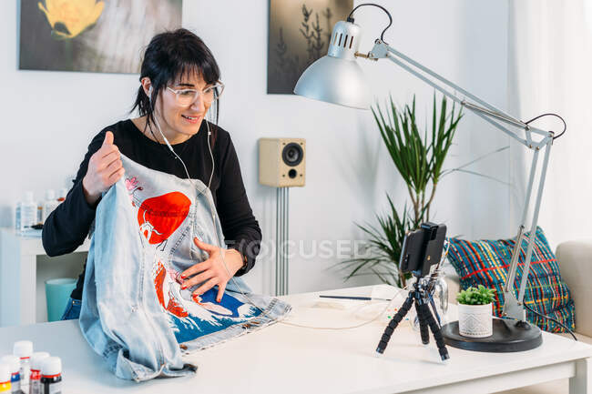 Vista lateral de la experta diseñadora femenina escuchando música en auriculares y dibujando boceto en chaqueta de mezclilla en la mesa en casa - foto de stock