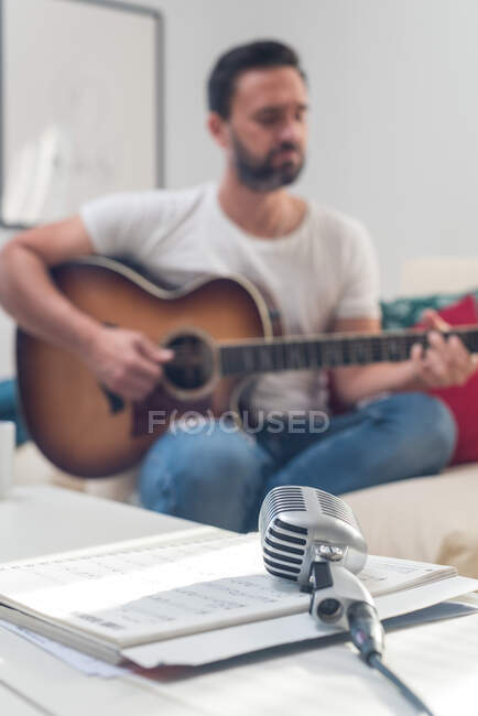 Винтажный микрофон, установленный на столе возле урожая, неузнаваемый бородатый мужчина-музыкант, играющий на акустической гитаре на диване — стоковое фото