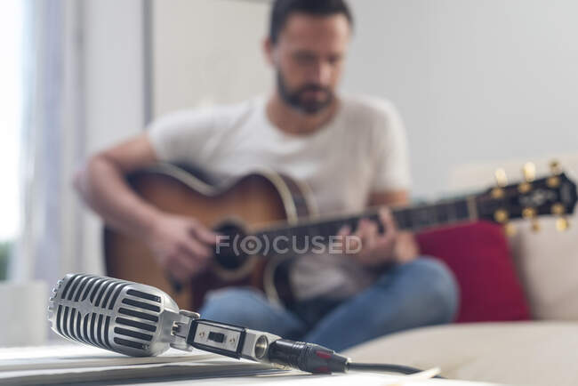 Вінтажний мікрофон, розміщений на столі поблизу врожаю невідомий бородатий чоловічий музикант грає на акустичній гітарі на дивані — стокове фото