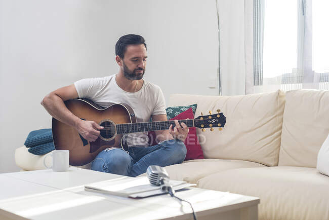 Colpo di barbuto musicista di sesso maschile che suona la chitarra acustica sul divano vicino al microfono vintage posto sul tavolo — Foto stock