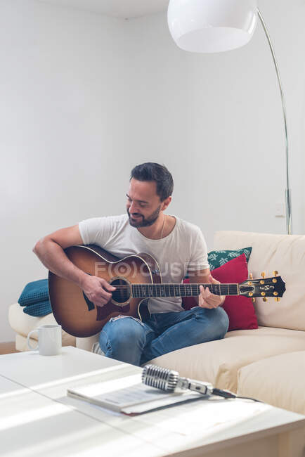 Урожай бородатого музыканта, играющего на акустической гитаре на диване рядом со старинным микрофоном, поставленным на стол — стоковое фото