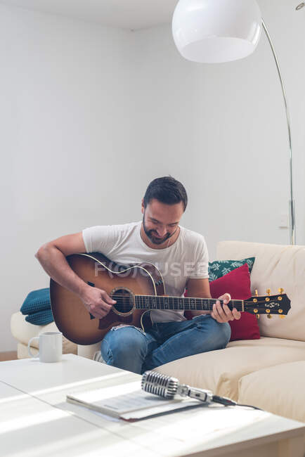 Урожай бородатого чоловіка-музиканта, який грає на акустичній гітарі на дивані біля старовинного мікрофона, розміщеного на столі — стокове фото