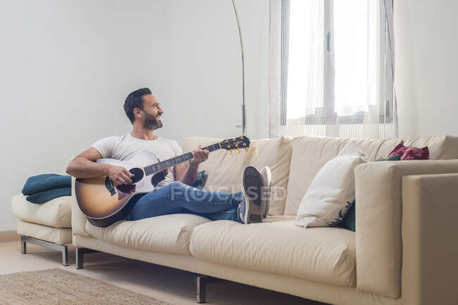 Все тело счастливого взрослого этнического музыканта в повседневной одежде расслабляется на удобном диване и играет на акустической гитаре дома — стоковое фото