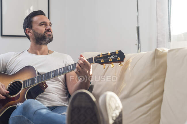 Все тело взрослого этнического мужчины-музыканта в повседневной одежде расслабляется на удобном диване и играет на акустической гитаре дома — стоковое фото