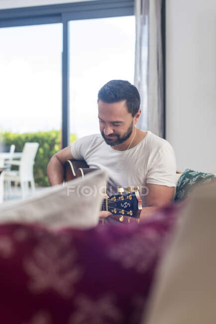 Colpo di barbuto musicista di sesso maschile suonare la chitarra acustica sul divano vicino alla finestra a casa — Foto stock