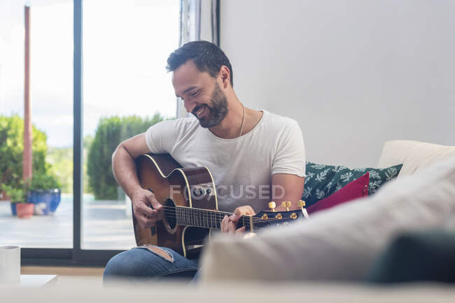 Бородатый музыкант играет на акустической гитаре на диване у окна дома — стоковое фото
