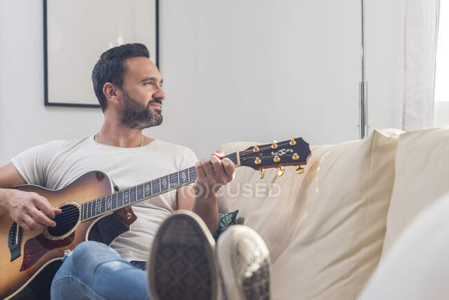 Corps complet de musicien masculin ethnique adulte en vêtements décontractés relaxant sur un canapé confortable et jouant de la guitare acoustique à la maison — Photo de stock