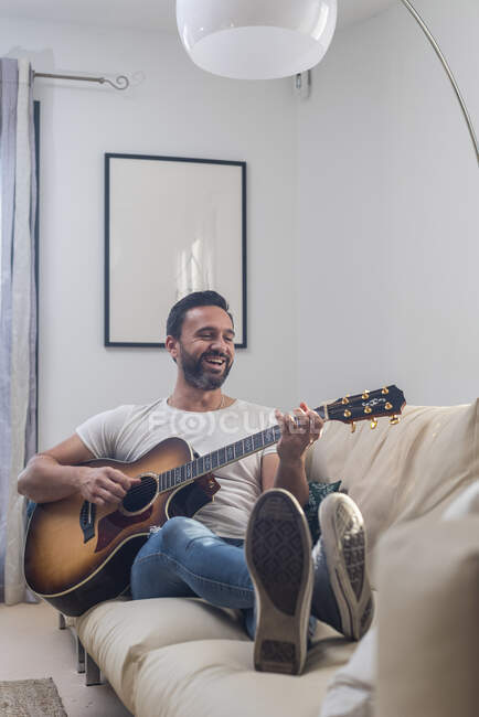Full body of happy adult ethnic male musician in casual clothes relaxing on comfortable sofa and playing acoustic guitar at home — Stock Photo