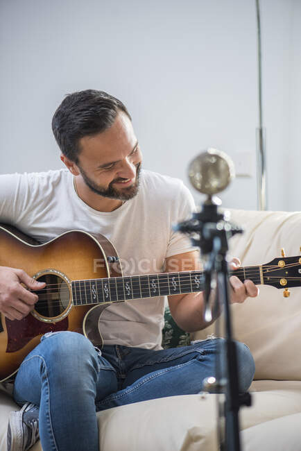 Урожай бородатого чоловіка-музиканта, який грає на акустичній гітарі на дивані біля старовинного мікрофона — стокове фото