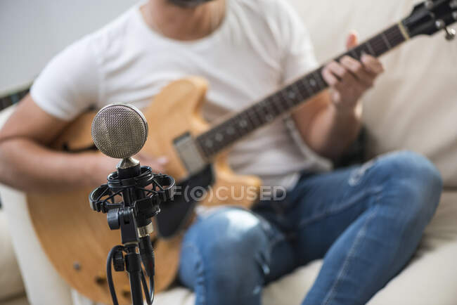 Micrófono vintage cerca de cultivo irreconocible barbudo músico masculino tocando la guitarra acústica en el sofá - foto de stock