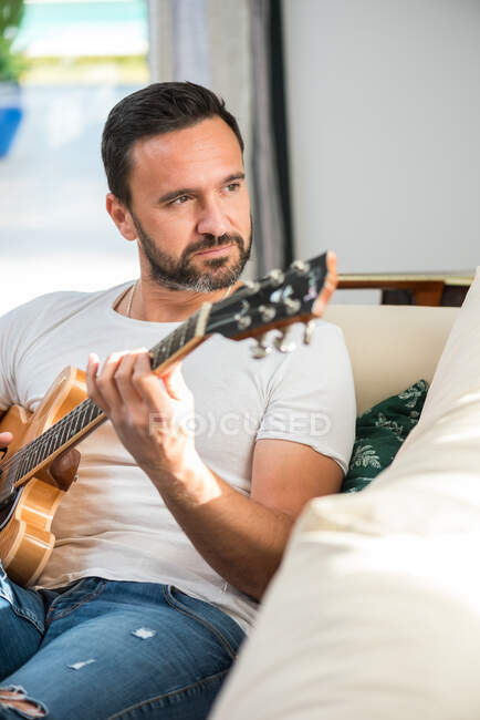 Дорослий етнічний чоловічий музикант в повсякденному одязі розслабляється на зручному дивані і грає на акустичній гітарі вдома — стокове фото
