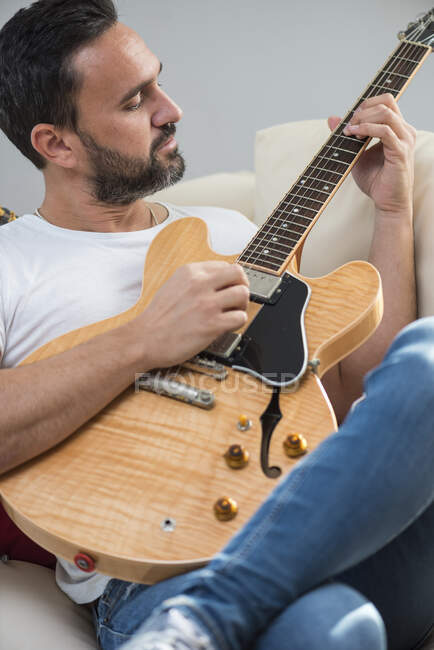 Взрослый мужчина-музыкант в повседневной одежде расслабляется на удобном диване и играет на акустической гитаре дома — стоковое фото