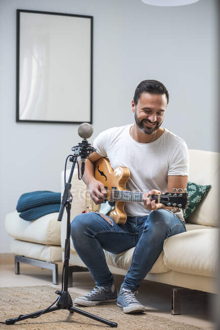 Alegre adulto hispano barbudo músico masculino en ropa casual que pone la guitarra eléctrica y canta sentado en un sofá suave en casa - foto de stock