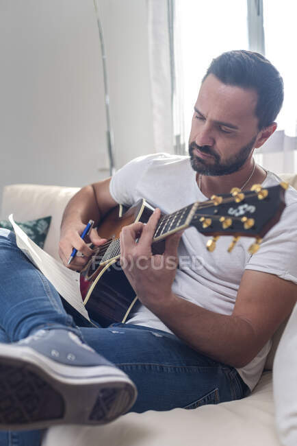 Концентрированный молодой бородатый этнический музыкант в повседневной одежде пишет на нотной бумаге, сочиняя песню, сидя на диване с акустической гитарой — стоковое фото
