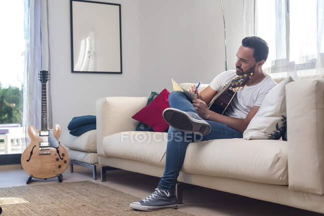 Full body of concentrated young bearded ethnic male musician in casual clothes writing on music paper while composing song sitting on sofa with acoustic guitar — Stock Photo