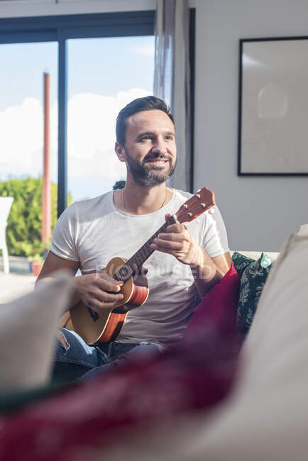 Позитивний бородатий етнічний гітарист посміхається, граючи укулеле сидячи на дивані в сучасній квартирі — стокове фото