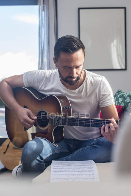 Músico masculino étnico adulto enfocado leyendo notas mientras toca la guitarra acústica sentado en el sofá en casa - foto de stock
