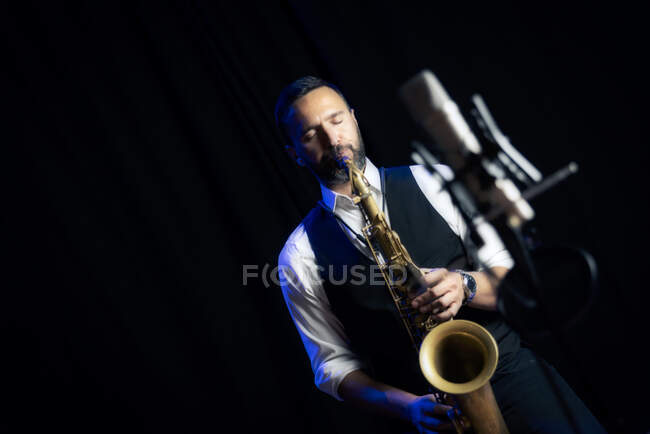 Урожай чоловічого музиканта в стильному вбранні, що стоїть біля мікрофона і грає на альт-саксофоні під час джазового концерту — стокове фото