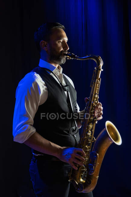 Vista lateral do músico masculino barbudo concentrado em roupa elegante tocando saxofone enquanto estava no palco durante o concerto — Fotografia de Stock