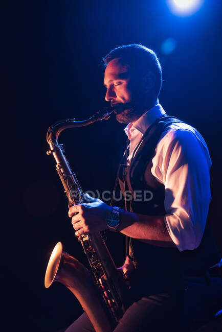Side view of concentrated bearded male musician in elegant outfit playing saxophone while standing on stage during concert — Stock Photo