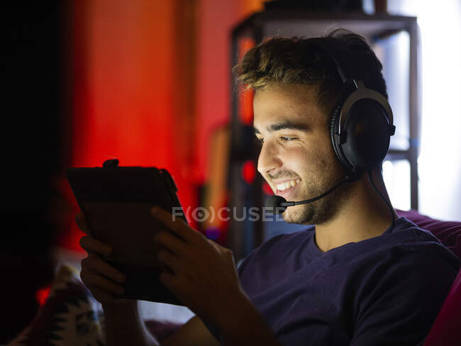 Contenuto giovane maschio in abbigliamento casual e cuffie wireless che gioca ai videogiochi su tablet mentre riposa sul divano in camera oscura — Foto stock