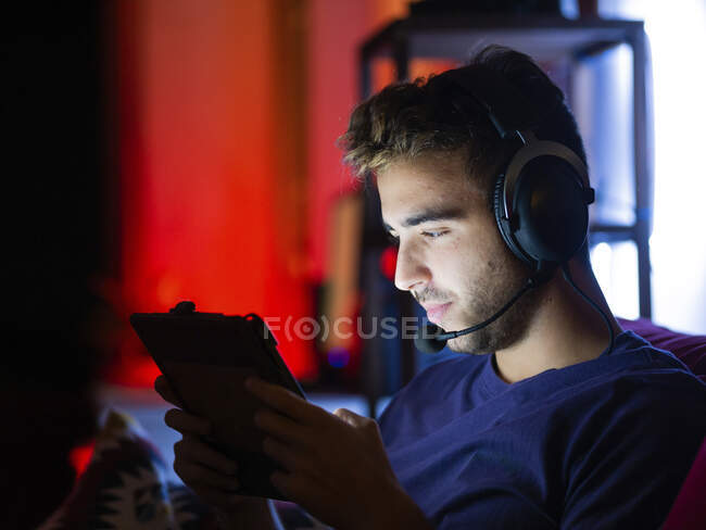 Zufriedener junger Mann in Freizeitkleidung und drahtlosen Kopfhörern, der Videospiele auf dem Tablet spielt, während er im dunklen Raum auf dem Sofa ruht — Stockfoto