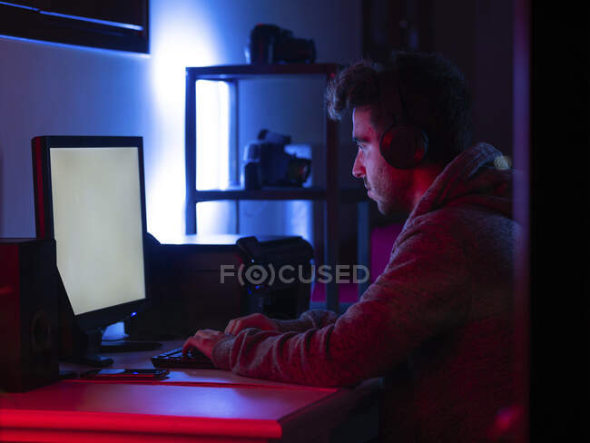 Vue de côté jeune brunet calme en tenue décontractée et casque sans fil naviguant sur l'ordinateur contemporain avec écran blanc tout en étant assis à table dans la pièce sombre — Photo de stock