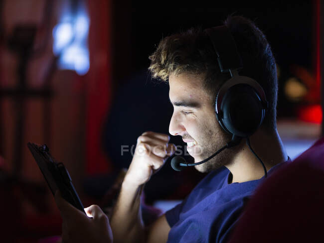 Side view joyful male in headphones browsing modern tablet and raising fist in celebration while sitting in dark room — Stock Photo
