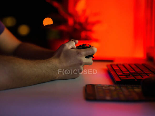 Recorte jugador masculino irreconocible usando gamepad y jugando videojuego mientras está sentado en la mesa con el ordenador en la habitación oscura - foto de stock