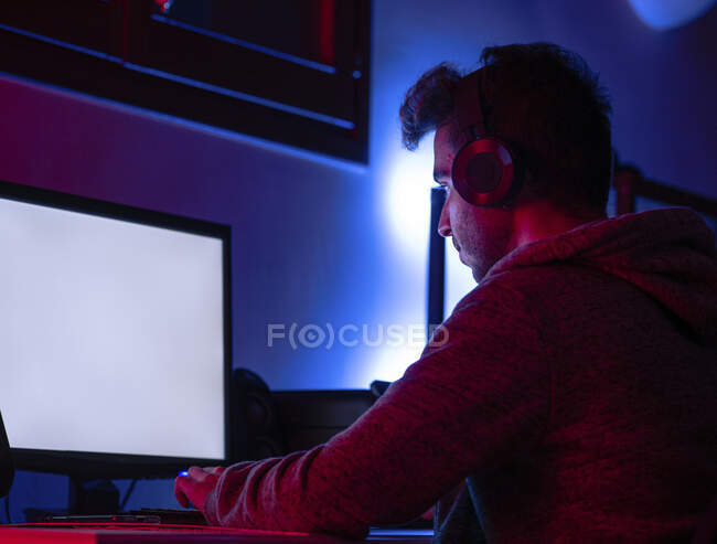 Вид сбоку успокаивает молодого брюнета в повседневной одежде и беспроводной гарнитуре, просматривающего современный компьютер с Blank-экраном, сидя за столом в темной комнате — стоковое фото