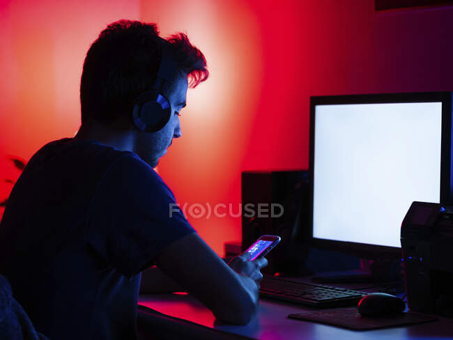 Side view focused young male in casual shirt and wireless headphones browsing mobile phone and working on computer with empty screen while sitting at desk in dark room — Stock Photo