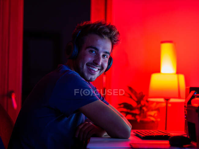 Vue latérale beau mâle heureux portant chemise décontractée et écouteurs sans fil penché sur le bureau avec ordinateur et regardant la caméra avec sourire dans la pièce sombre — Photo de stock