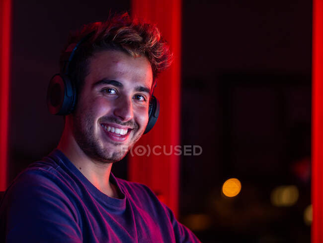 Guapo hombre feliz con camisa casual y auriculares inalámbricos apoyados en el escritorio con el ordenador y mirando a la cámara con sonrisa en la habitación oscura - foto de stock