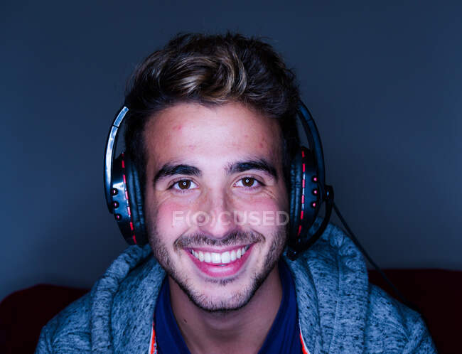 Vista frontal guapo hombre feliz con camisa casual y auriculares inalámbricos apoyados en el escritorio con el ordenador y mirando a la cámara con sonrisa en habitación oscura - foto de stock
