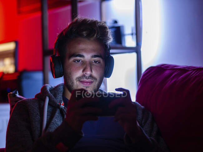 Zufriedener junger Mann in Freizeitkleidung und kabellosen Kopfhörern surft im Handy, während er sich im dunklen Raum auf dem Sofa ausruht — Stockfoto