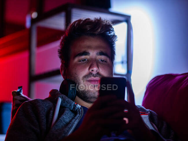 Contenuto giovane maschio in abbigliamento casual e cuffie wireless che naviga sul cellulare mentre riposa sul divano in camera oscura — Foto stock