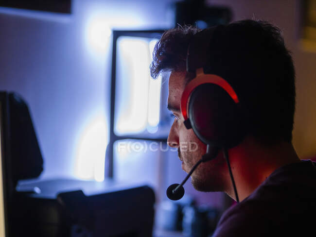 Side view handsome happy male wearing casual shirt and wireless headphones leaning on desk with computer and looking at screen with smile in dark room — Stock Photo
