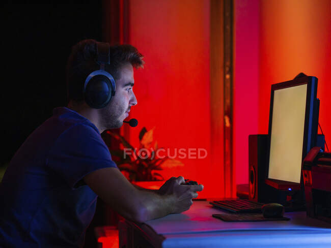 Side view calm young brunet in casual wear and wireless headset browsing contemporary computer with blank screen while sitting at table in dark room — Stock Photo