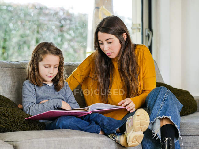 Content female teen looking the textbook page while spending time with sibling on couch at home — Stock Photo